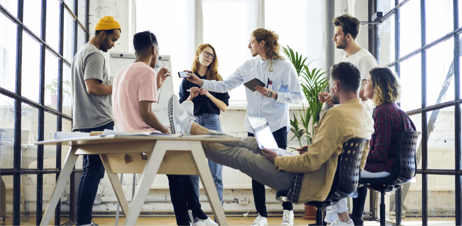 Colleagues, some seated and some standing around a table, discussing in front of a whiteboard